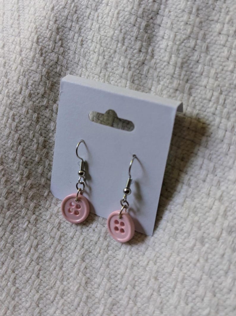 May include: A pair of pink button earrings with silver-colored hooks and chain attachments. The earrings are displayed on a white card with a small cutout at the top, set against a textured white background.