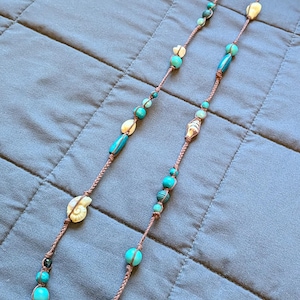May include: A long necklace featuring a brown braided cord accented with turquoise-colored beads in various shapes and sizes, interspersed with small white seashells. The design includes round, cylindrical, and textured beads along the cord.