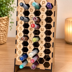 Peut inclure: Un organisateur de bureau en bois brun clair avec un motif en nid d'abeille composé de fentes hexagonales. Le panneau avant est gravé avec les chiffres 1 à 9 et les lettres minuscules a à z. Plusieurs marqueurs colorés sont insérés.