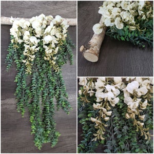 May include: A white floral garland with green leaves hanging from a branch. The garland is made of artificial flowers and leaves and is perfect for adding a touch of greenery to any space.