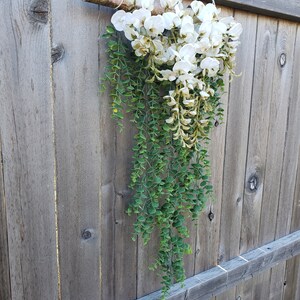 May include: A white and green floral garland hanging from a wooden branch. The garland is made of white flowers and green leaves.