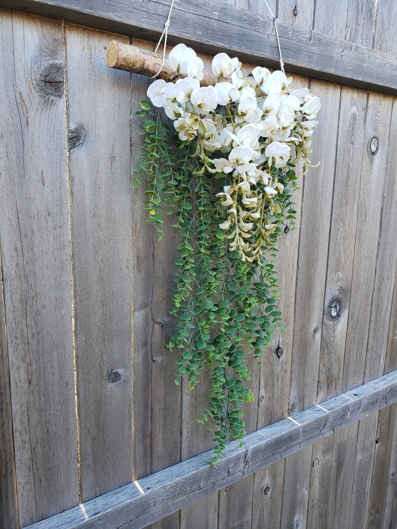 May include: A white and green floral garland hanging from a wooden branch. The garland is made of white flowers and green leaves.