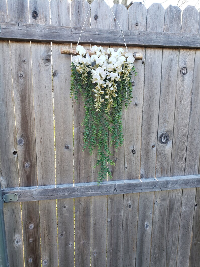 May include: A white floral garland with green leaves hanging from a wooden branch. The garland is attached to a wooden fence.