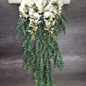 May include: A white floral garland with green leaves hanging from a piece of driftwood. The garland is made of artificial flowers and leaves.