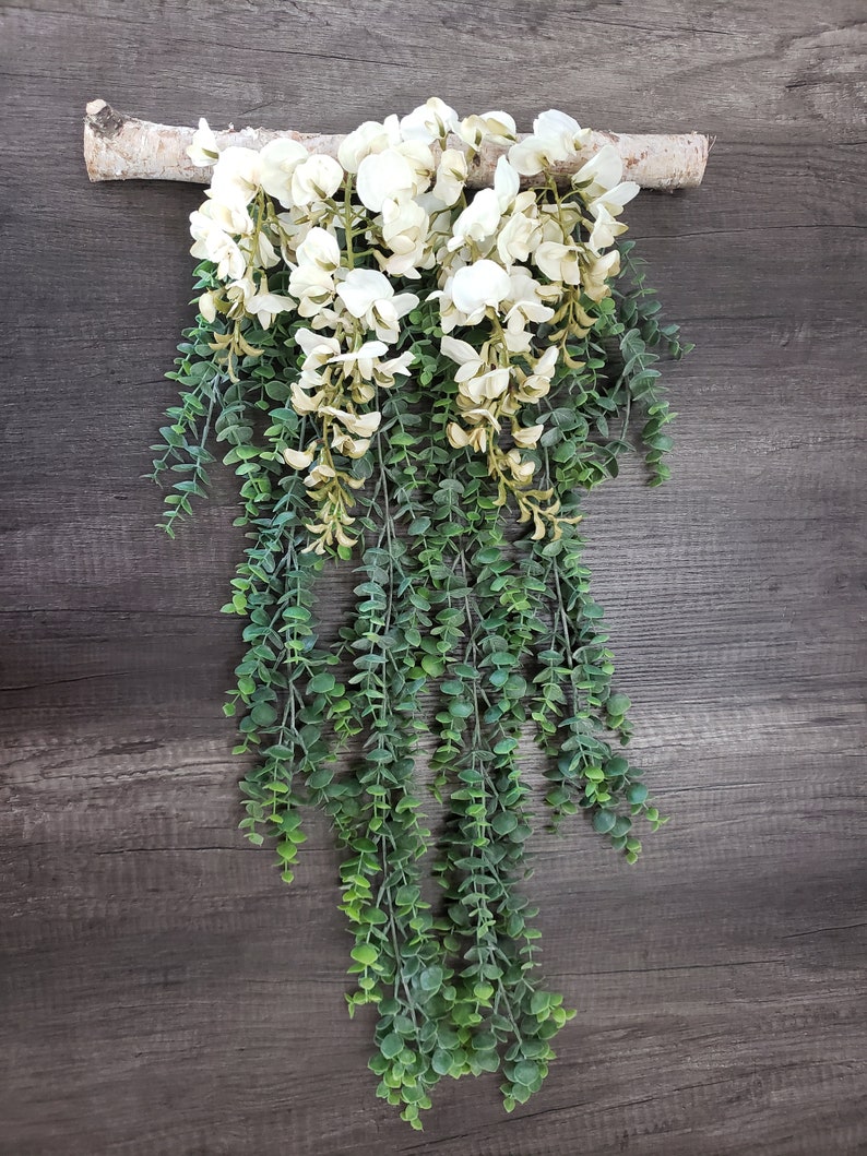May include: A white floral garland with green leaves hanging from a piece of driftwood. The garland is made of artificial flowers and leaves.