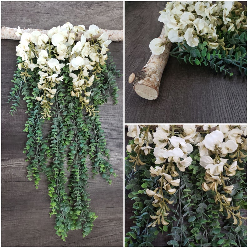 May include: A white floral garland with green leaves hanging from a branch. The garland is made of artificial flowers and leaves and is perfect for adding a touch of greenery to any space.