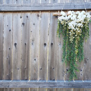 May include: A white and green floral garland hanging from a wooden branch against a weathered wooden fence.