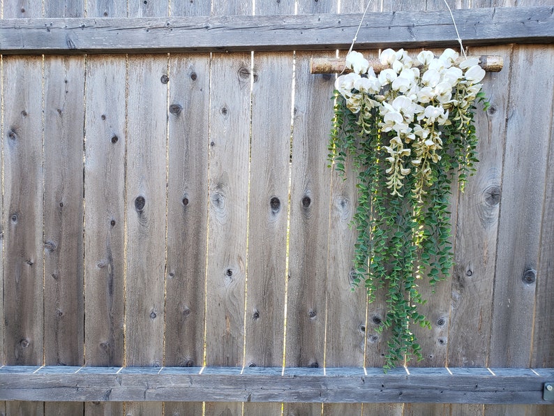 May include: A white and green floral garland hanging from a wooden branch against a weathered wooden fence.