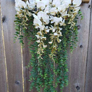 May include: A white flower garland with green leaves hanging from a wooden branch. The garland is made of artificial flowers and leaves.