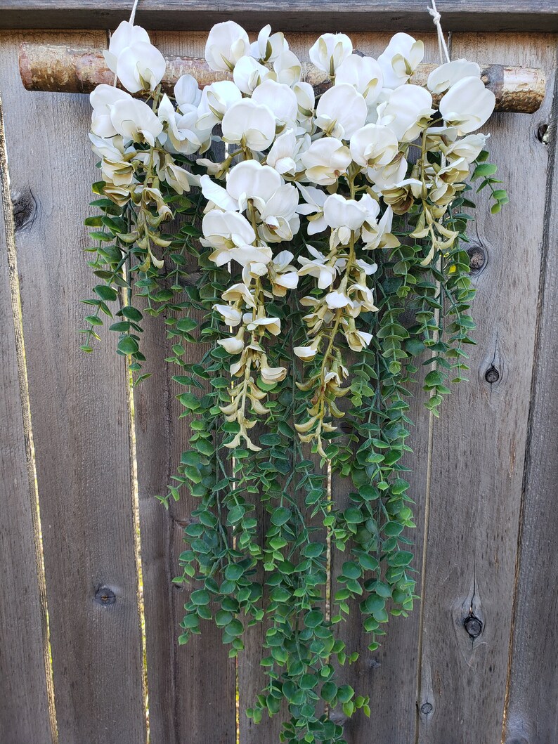 May include: A white flower garland with green leaves hanging from a wooden branch. The garland is made of artificial flowers and leaves.