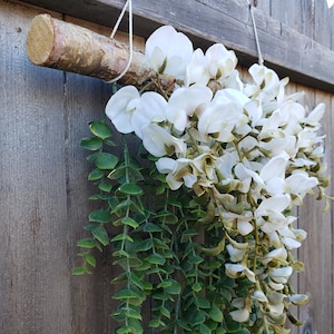 May include: A white floral garland with green leaves hanging from a wooden branch. The garland is attached to a wooden fence.