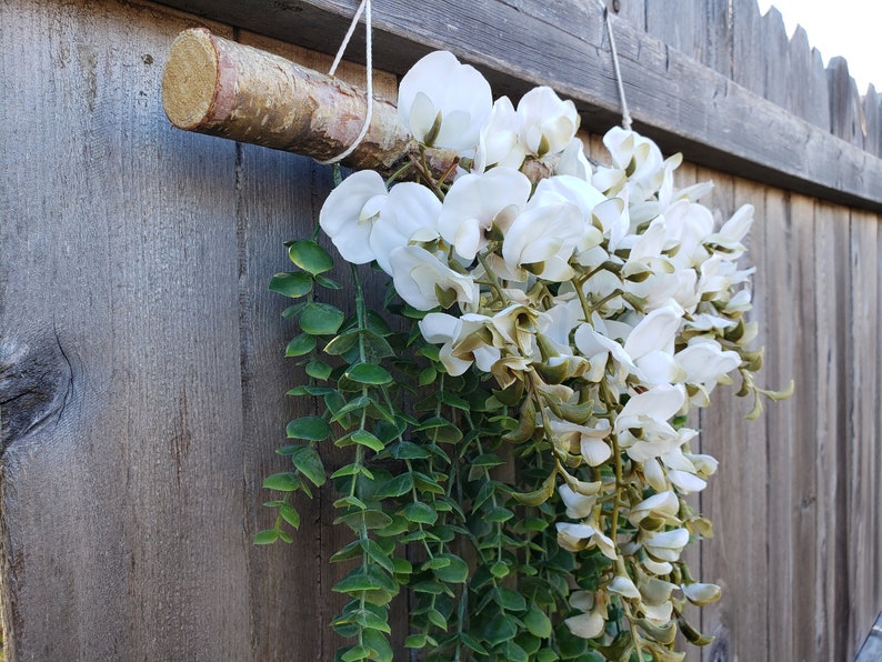 May include: A white floral garland with green leaves hanging from a wooden branch. The garland is attached to a wooden fence.