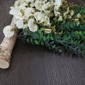 May include: A white and green floral arrangement with white flowers and green leaves on a brown wooden surface.