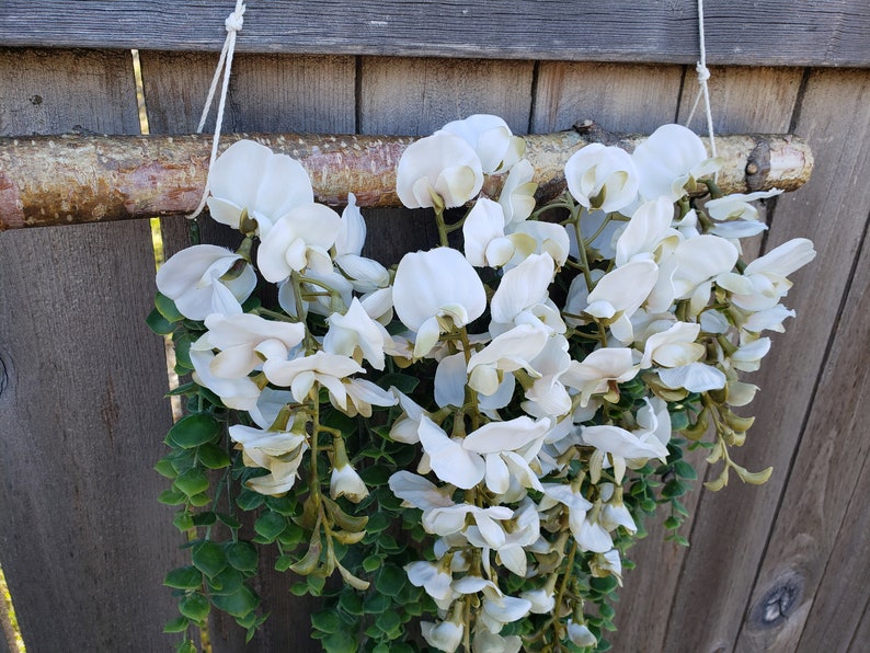 May include: A white floral garland with green leaves hanging from a wooden branch. The garland is made of artificial flowers and is perfect for decorating a home or event.