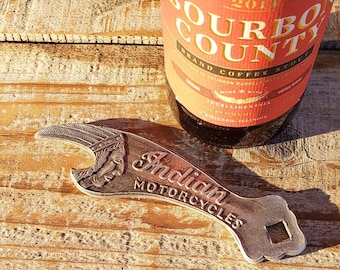 May include: A silver bottle opener with a stylized Indian motorcycle logo and the words "Indian Motorcycles". The bottle opener is sitting on a wooden surface next to a bottle of Goose Island Bourbon County Brand Coffee Stout.
