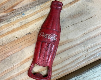May include: Red metal bottle opener shaped like a Coca-Cola bottle. The bottle opener has a white Coca-Cola logo.