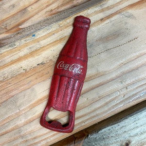 Puede incluir: Abrebotellas de metal rojo con forma de botella de Coca-Cola. El abrebotellas tiene un logotipo de Coca-Cola blanco.