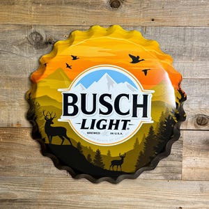 May include: A large, metal Busch Light bottle cap sign. The sign features a scenic landscape with deer, trees, and birds against a sunset sky. The Busch Light logo is prominently displayed in the center.
