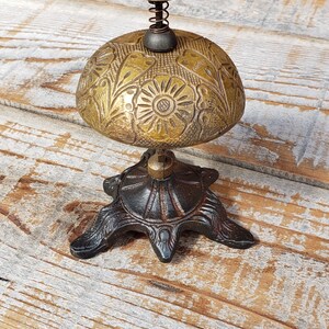 May include: Antique brass bell with floral detailing. The bell has a domed shape with a spring and a small knob on top. It sits on a decorative, dark metal base with three feet. The bell is displayed on a rustic wooden surface.