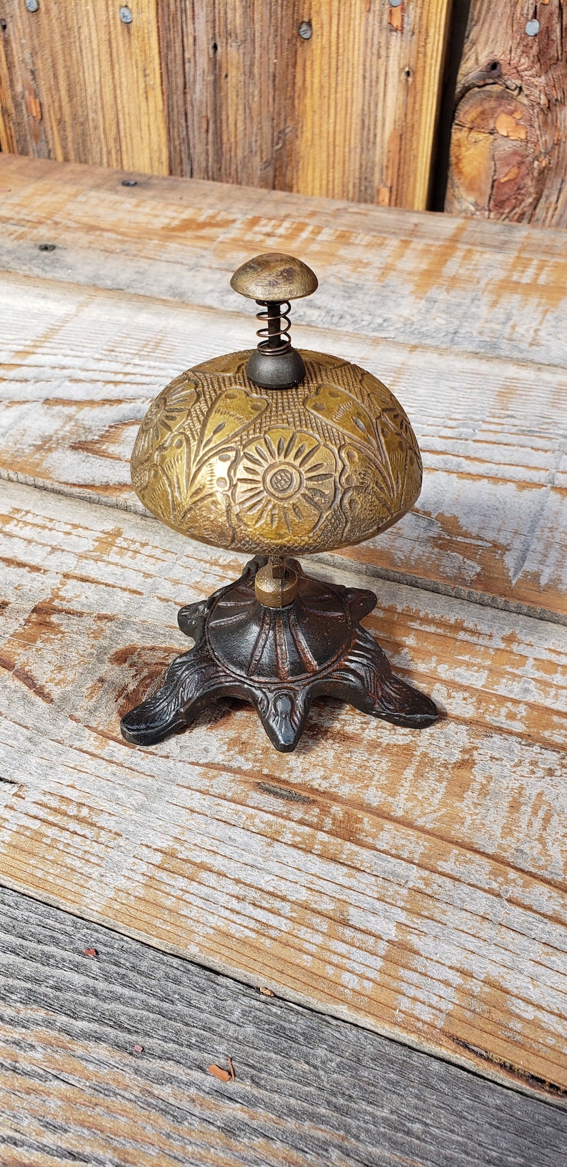 May include: Antique brass bell with floral detailing. The bell has a domed shape with a spring and a small knob on top. It sits on a decorative, dark metal base with three feet. The bell is displayed on a rustic wooden surface.
