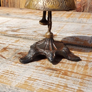 May include: An antique brass bell with a floral design on the bell and a spring-loaded clapper. The bell is mounted on a decorative, dark metal base with three feet. The background is a weathered wooden surface.