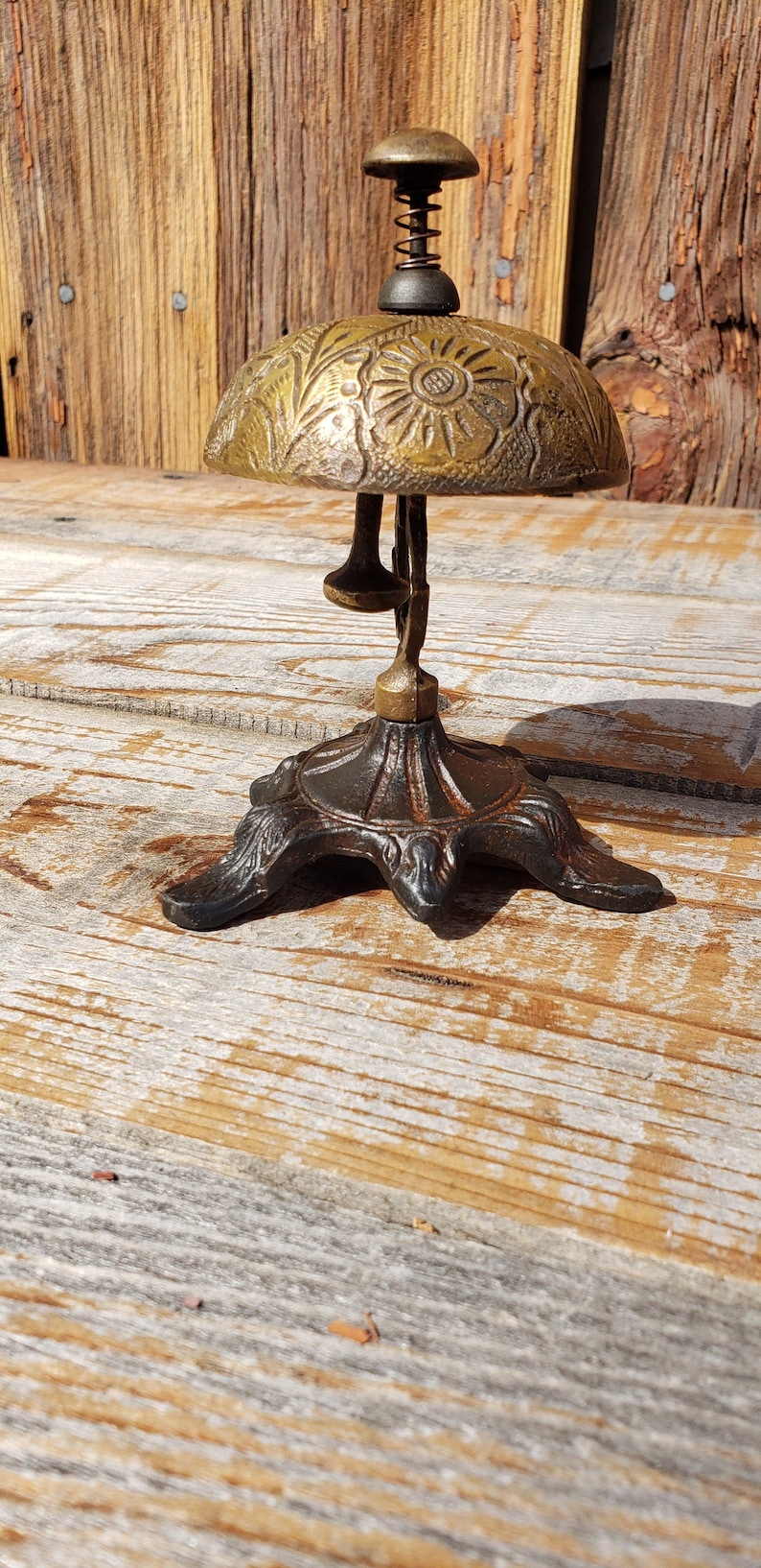 May include: An antique brass bell with a floral design on the bell and a spring-loaded clapper. The bell is mounted on a decorative, dark metal base with three feet. The background is a weathered wooden surface.