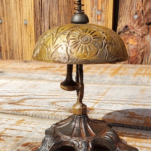 May include: Antique brass bell with a floral design on the dome. The bell has a spring-loaded mechanism and a decorative base. The bell is made of brass and has a vintage look. The bell is a decorative item.