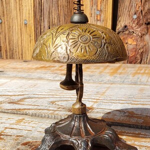 May include: Antique brass bell with a floral design on the dome. The bell has a spring-loaded mechanism and a decorative base. The bell is made of brass and has a dark, aged patina. The bell is a vintage item.