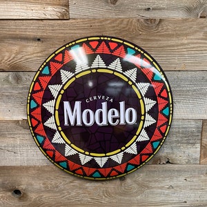 May include: A round, metal sign with a black background and a colourful, geometric design. The sign features the text "Cerveza Modelo" in white letters.
