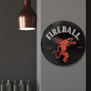 以下が含まれることがあります： 「Fireball Red Hot」の文字と、炎をまとった赤い悪魔のキャラクターが描かれた、黒と赤の丸い金属製の看板です。