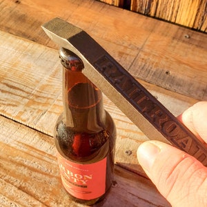 May include: A brown bottle opener with the word "Railroad" embossed on it is being used to open a brown glass bottle of beer. The bottle has a red label with the text "Iron County Beer Company" on it.