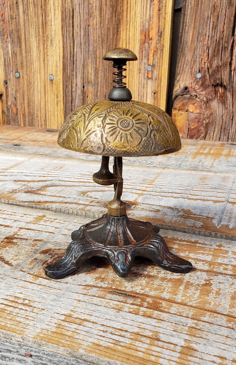 May include: An antique brass desk bell with a floral design on the dome. The bell has a spring and a small handle on top. The base is ornate and dark brown. The bell is set on a wooden surface.