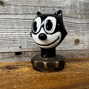 Könnte beinhalten: Schwarze und weiße Keramikfigur eines Comic-Katzen mit einem breiten Lächeln und großen Augen. Die Figur ist auf einem Messingsockel montiert.