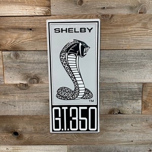 May include: A rectangular metal sign with the word "SHELBY" at the top, above a cobra emblem. Below the emblem is the text "G.T.350". The sign has a silver background and a black and white cobra design. The sign is mounted on a wooden surface.