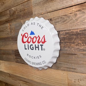Peut inclure: Un grand panneau métallique blanc en forme de capsule de bouteille. Il présente le logo Coors Light en rouge et bleu, avec les mots "COLD AS THE" et "ROCKIES" en blanc. Le panneau est fixé sur un fond en bois.