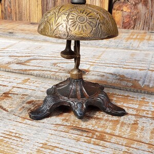 May include: Antique brass bell with a floral design on the dome. The bell has a spring and a clapper, and sits on a decorative, dark metal base. The background is a rustic wooden surface.
