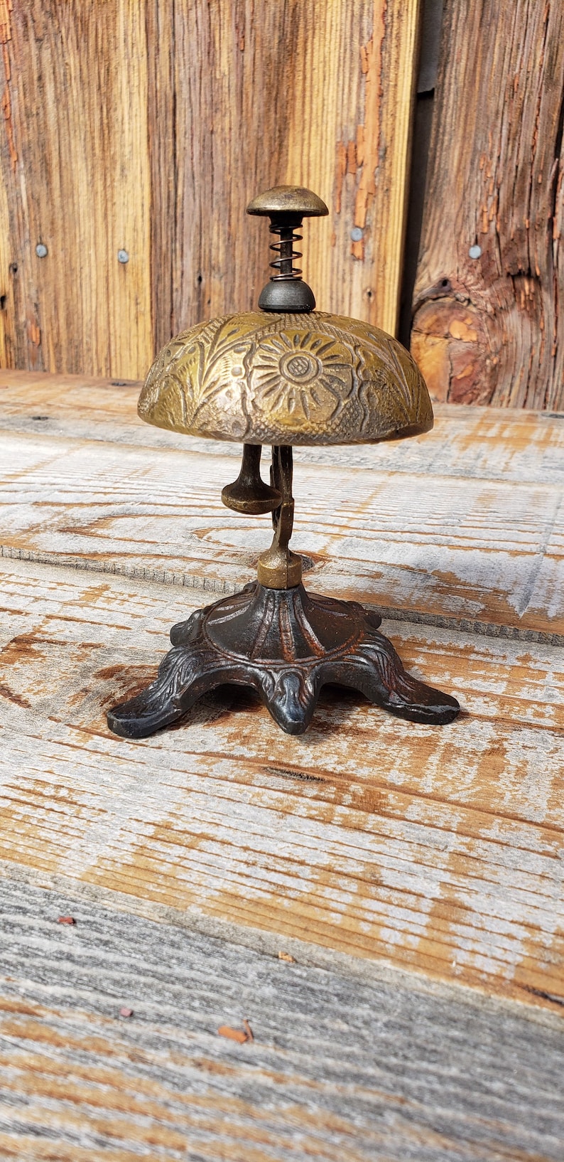 May include: Antique brass bell with a floral design on the dome. The bell has a spring and a clapper, and sits on a decorative, dark metal base. The background is a rustic wooden surface.