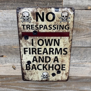 May include: A rectangular metal sign with a distressed finish, featuring the text "NO TRESPASSING" above a red line, and "I OWN FIREARMS AND A BACKHOE" below. The sign includes skull and crossbones graphics and bullet hole details.