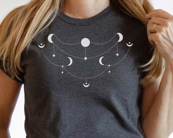 May include: Dark gray t-shirt featuring a white celestial design of moons and stars. The graphic is a series of crescent moons, a full moon, and stars connected by dotted lines. The shirt is a crew neck and is paired with blue jeans.