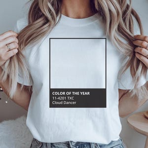 May include: White t-shirt with the text "COLOR OF THE YEAR" and the color code 11-4201 TXC, "Cloud Dancer" in a black rectangle. The model is wearing the shirt and holding her hair. The shirt is a casual style.