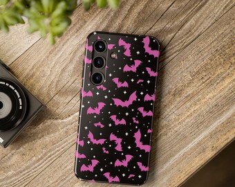 May include: Black smartphone case featuring a pattern of pink bats and white stars. The case is on a wooden surface, with a camera and plant in the background. The case has three camera lenses.