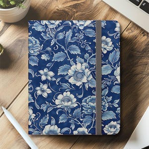 Peut inclure: Un carnet bleu avec un motif floral. La couverture présente un motif de fleurs et de feuilles blanches et bleu clair sur un fond bleu foncé. Une bande élastique grise maintient le carnet fermé.