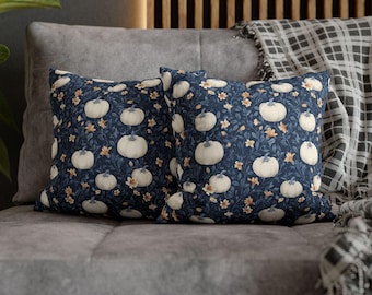May include: Two square throw pillows with a navy blue background and a repeating pattern of white pumpkins and small orange flowers. The pillows are on a gray couch with a plaid blanket.
