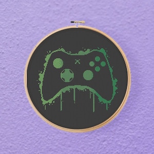 May include: A green and black video game controller in a circular embroidery hoop. The controller is outlined in green and has a black background.