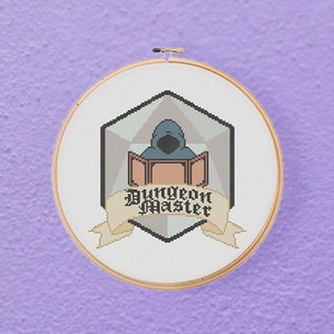 May include: A white cross-stitch embroidery hoop with a gray and brown design of a Dungeons and Dragons Dungeon Master icon. The icon features a hooded figure sitting behind a screen with a 20-sided die in the background.