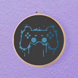 May include: A cross-stitch embroidery hoop featuring a black background with a blue controller design. The controller has a dripping paint effect. The wooden hoop is round and has a metal clasp. The background is a textured purple.