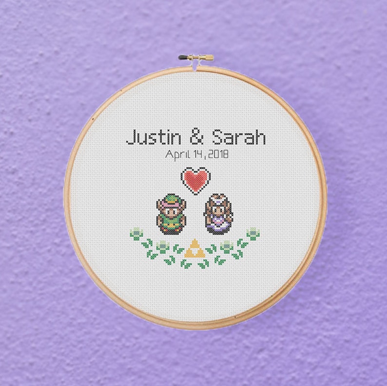 May include: A white embroidery hoop with a cross-stitch design featuring two pixelated characters from the Legend of Zelda video game series. The characters are standing next to each other with a heart above them. The text "Justin & Sarah April 14, 2018" is above the characters.