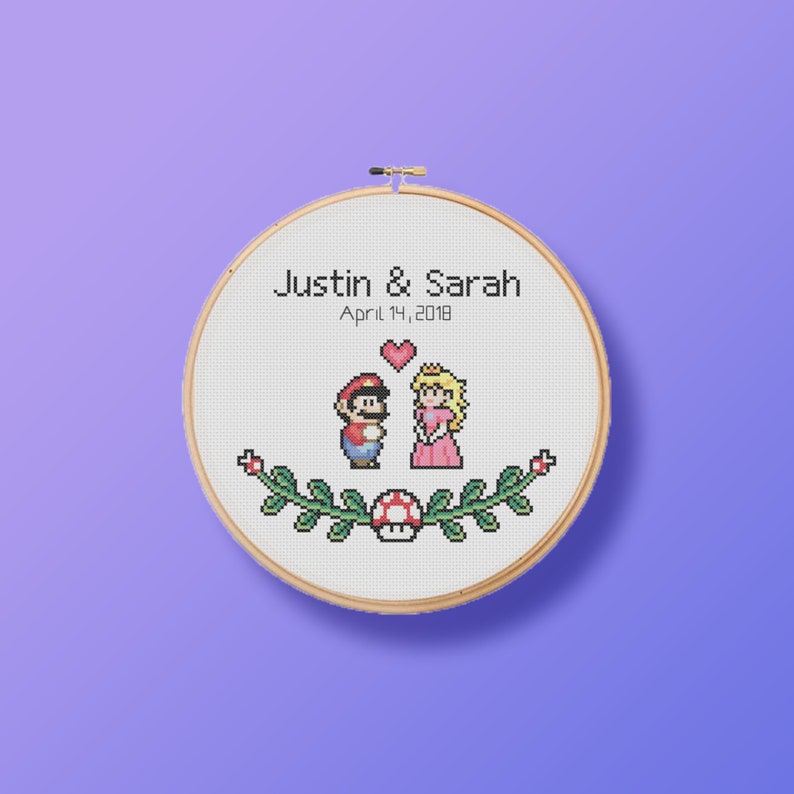 May include: A cross-stitch embroidery hoop with a pixelated image of Mario and Princess Peach from the Super Mario Bros. video game. The hoop is decorated with a green vine and a red mushroom. The text "Justin & Sarah April 14, 2018" is embroidered above the characters.