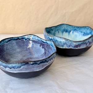 May include: Two ceramic bowls with irregular, wavy rims. The front bowl features a glossy purple interior transitioning to a dark blue and white drip glaze at the edge, with a matte black exterior. The back bowl has a light blue interior.
