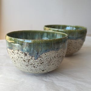May include: Two ceramic bowls featuring a speckled cream-coloured base with dark brown flecks. The upper portion of each bowl is finished with a glossy, dripping glaze in shades of deep forest green and muted blue, creating a smooth transition.